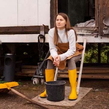 Woman in yellow boots sitting on a chair outdoors with a black bucket and shovel nearby.