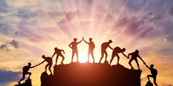Silhouettes of people helping each other climb a hill at sunset.