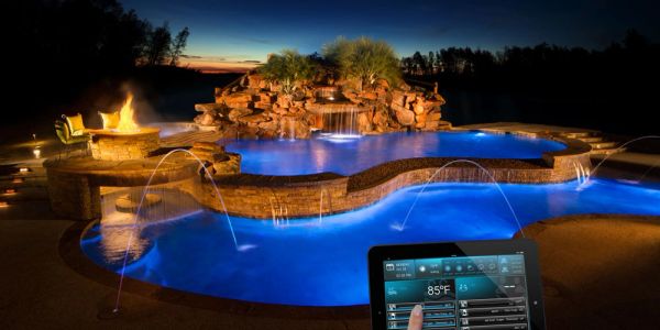 Luxury pool with lighting, waterfalls, and fire pit controlled by a tablet at night.
