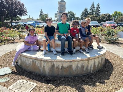Six children sitting on a circular concrete structure outdoors on a sunny day.