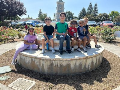 Six children sitting on a circular concrete structure outdoors on a sunny day.