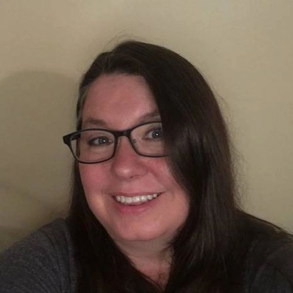Smiling woman with long dark hair and glasses against a plain background.