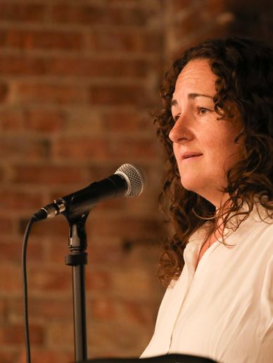 Woman speaking into a microphone against a brick wall background.