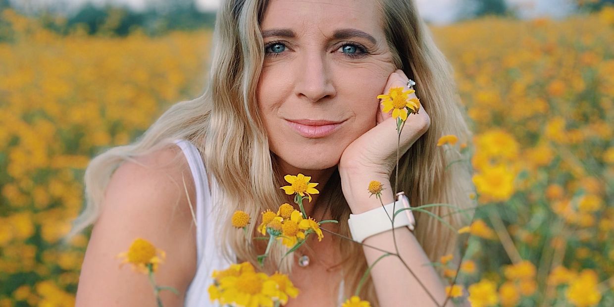 Woman with blue eyes amidst yellow flowers, resting her chin on her hand.