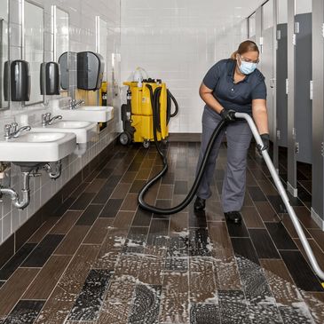 A janitor vacuuming a wet floor in a restroom while wearing a mask.