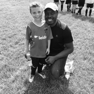 A man kneels next to a smiling boy in a soccer uniform on a grassy field.