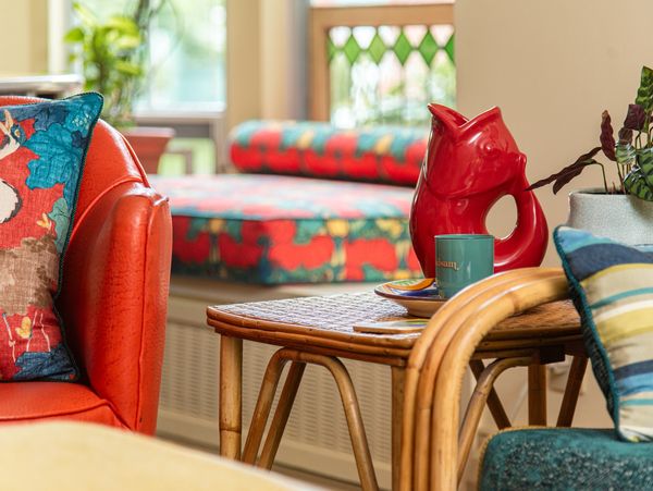 Colorful living room with patterned cushions and a red ceramic pitcher on a wicker table.