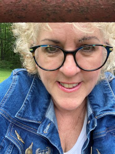Smiling woman with curly hair and glasses outdoors in a denim jacket.