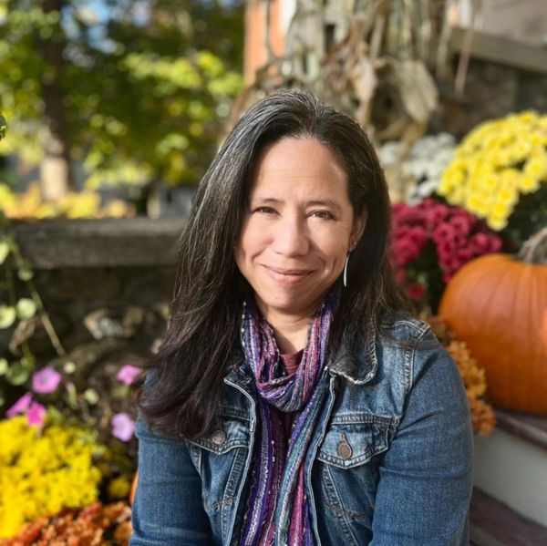 Woman with long dark hair smiling outdoors surrounded by colorful flowers and a pumpkin.