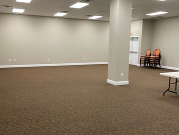 Large empty room with brown carpet and beige walls, lit by ceiling lights.