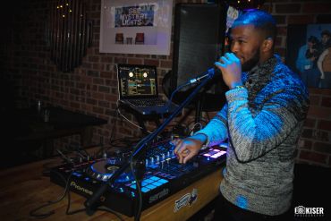 DJ performing with mixing console and laptop in a dimly lit room.