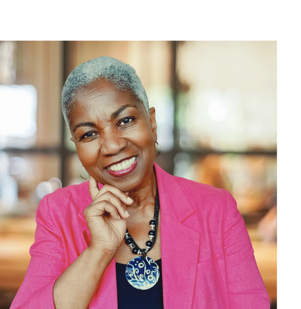 A lady in a pink jacket, smiling at a desk, with head leaned to one side and finger on cheek