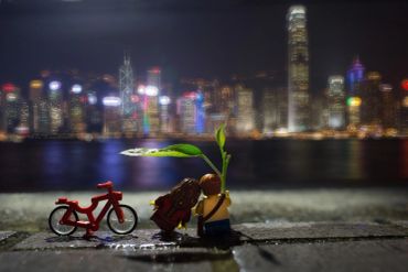 Miniature figures with a plant and bike by a waterfront city skyline.