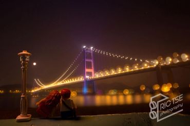 Night view of a lit suspension bridge with a person sitting and watching.