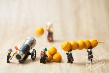 Miniature figures with a toy cannon and fried snacks on sticks.