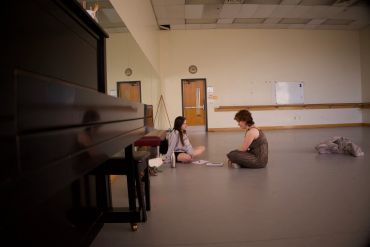 Two people sitting on a dance studio floor near a piano.
