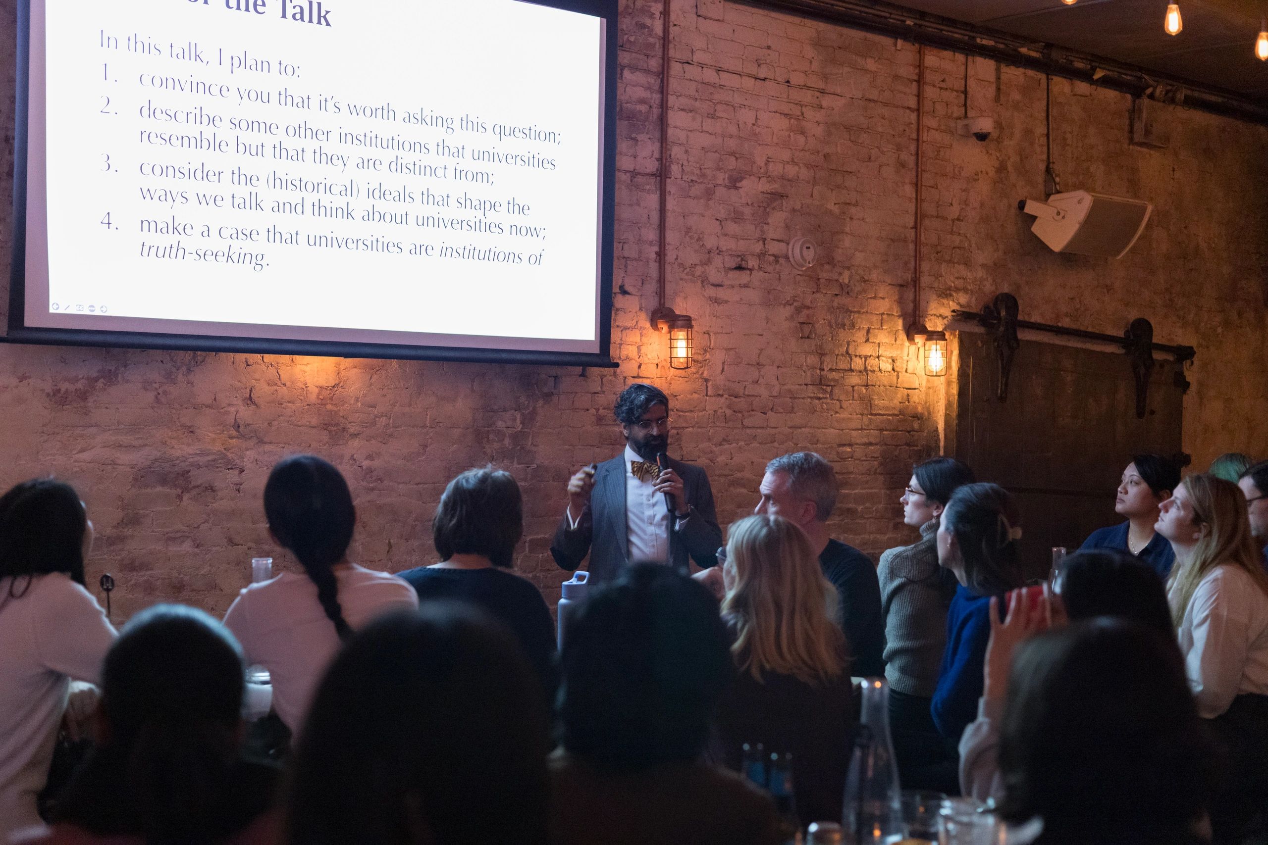A speaker presents a lecture to an attentive audience in a cozy, brick-walled venue.