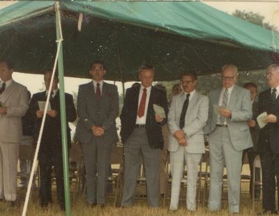 Group of men in suits standing under a green tent outdoors.