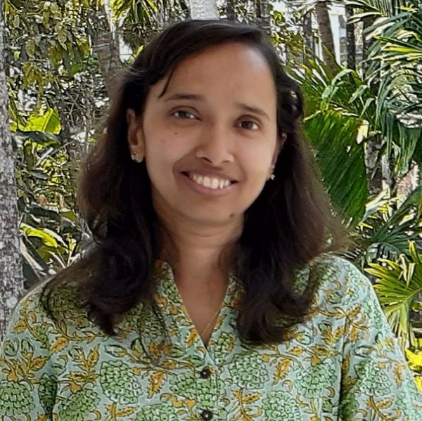 A smiling woman in a green floral top standing outdoors with lush greenery behind.