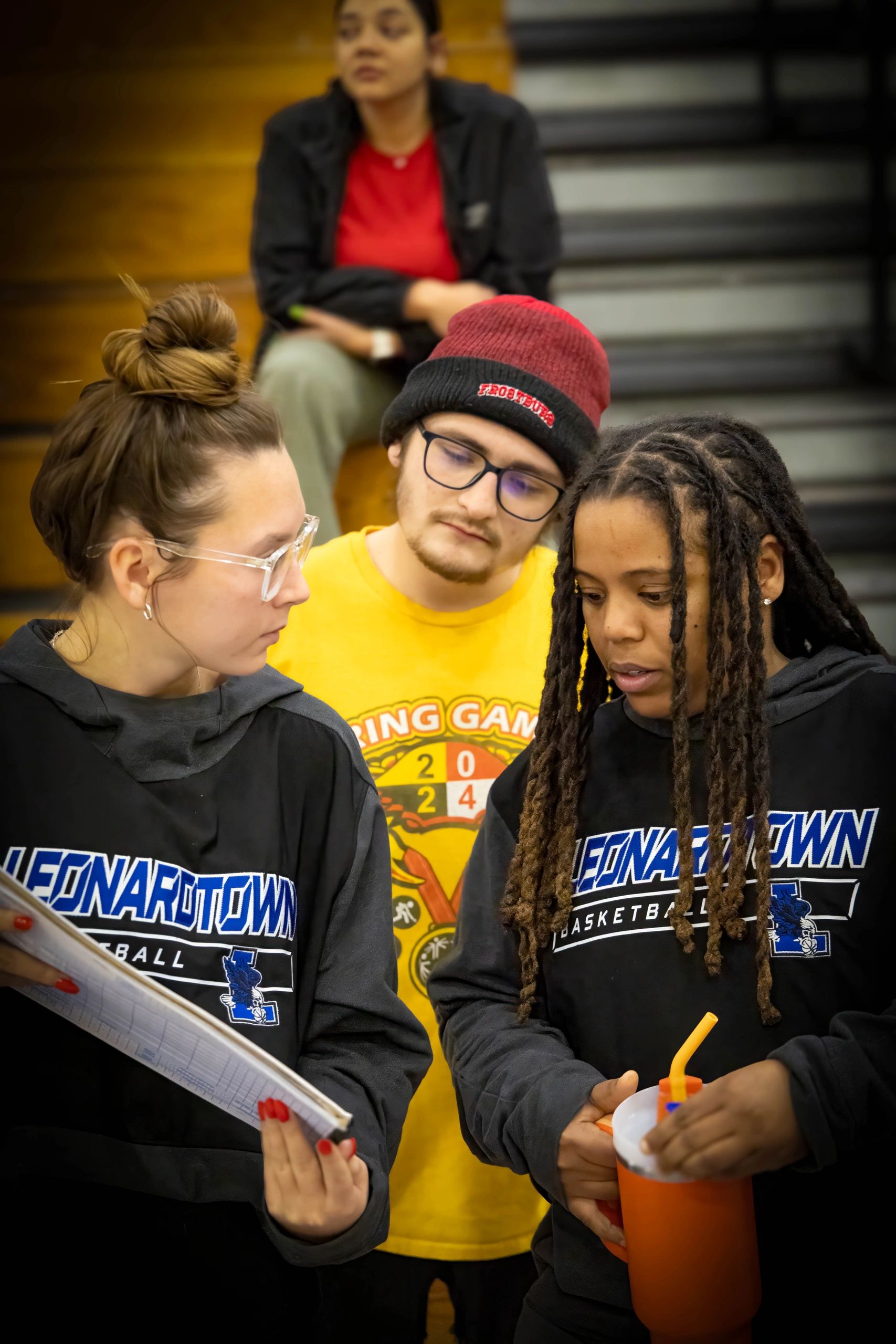 Three focused young people in Leonardtown basketball hoodies, one holding a clipboard and another an orange drink.