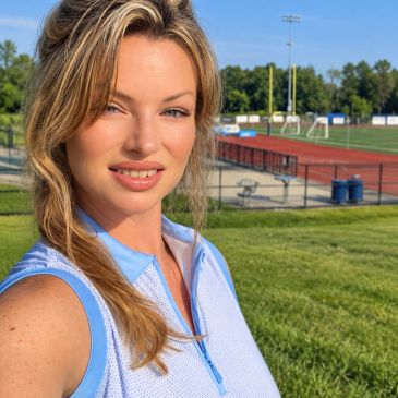 Smiling woman in a blue sleeveless top outdoors near a sports field.