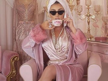 Elegant woman in pink robe and sunglasses enjoying tea in a luxurious pink room.