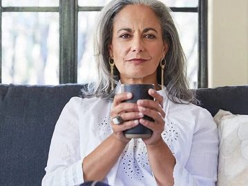A confident mature woman holding a black cup, sitting comfortably indoors.