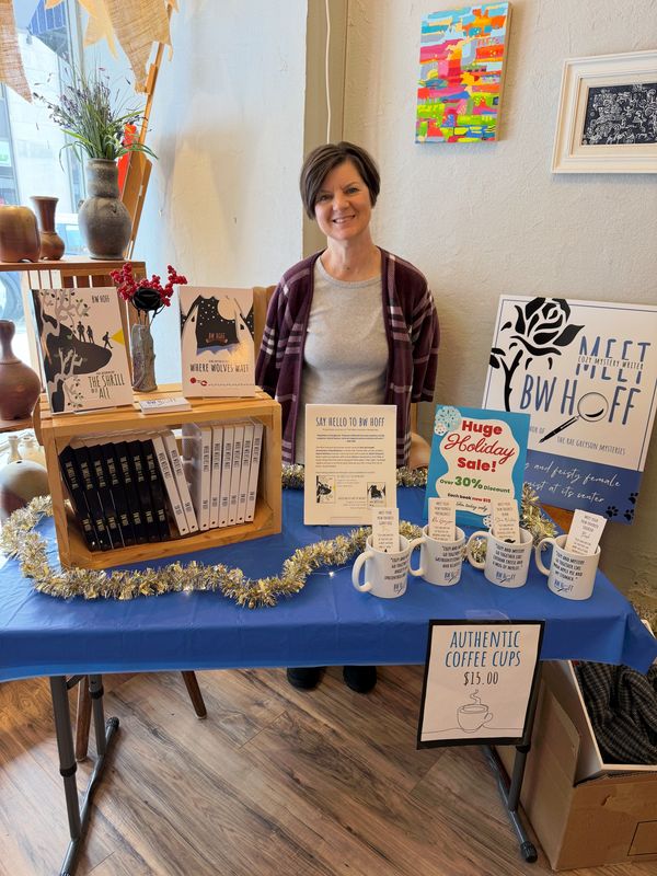 Woman smiling behind a holiday sale table with books and coffee mugs.