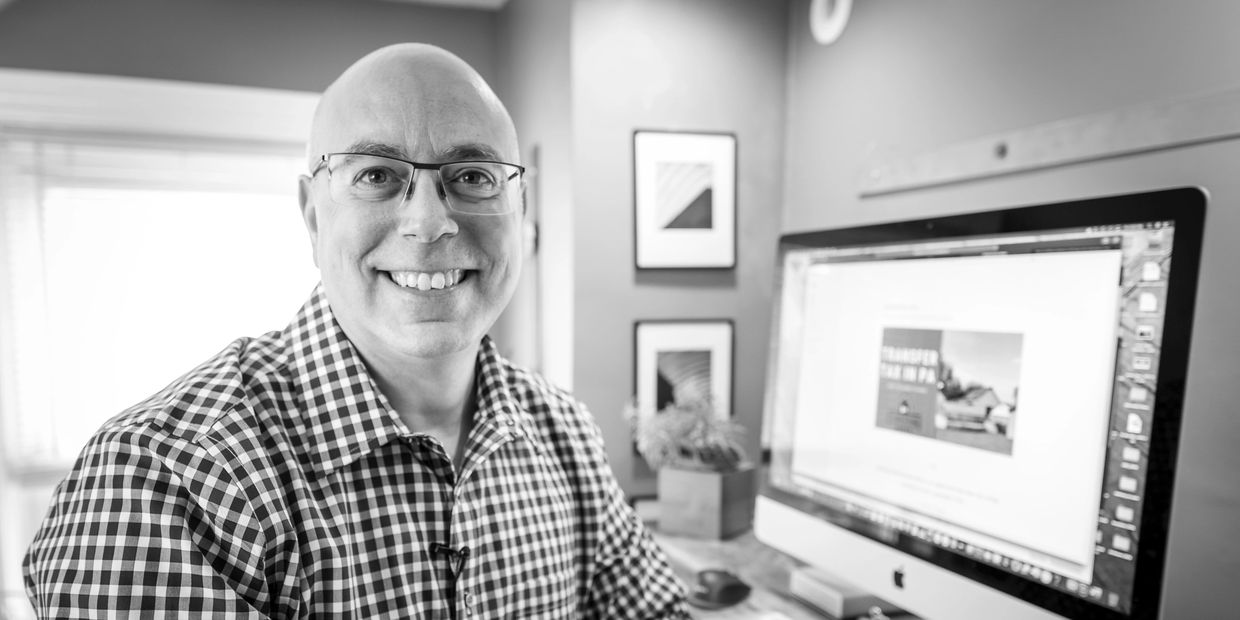A smiling man in glasses sits at a desk with a computer in a home office.