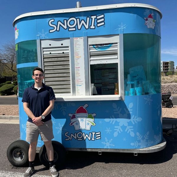 Young man standing in front of a blue Snowie shaved ice stand on a sunny day.