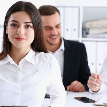Confident woman in white shirt with colleagues in office background.