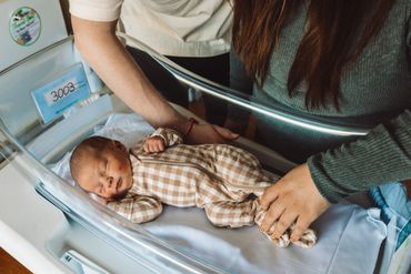 Newborn baby lying in a hospital bassinet with parents' hands nearby.
