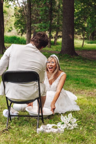 Bride joyfully washing groom's feet outdoors after wedding.