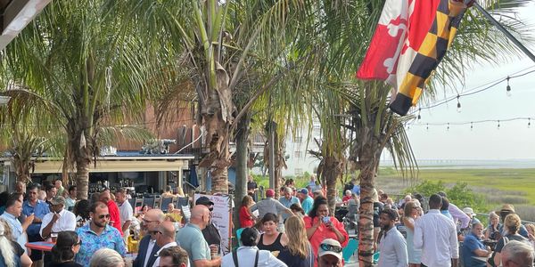 Outdoor social gathering under palm trees with Maryland flags and a marsh view.