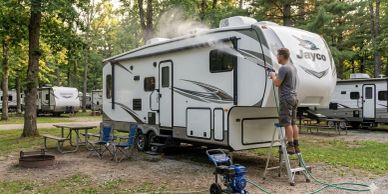 Man pressure washing a Jayco RV at a campsite surrounded by trees.