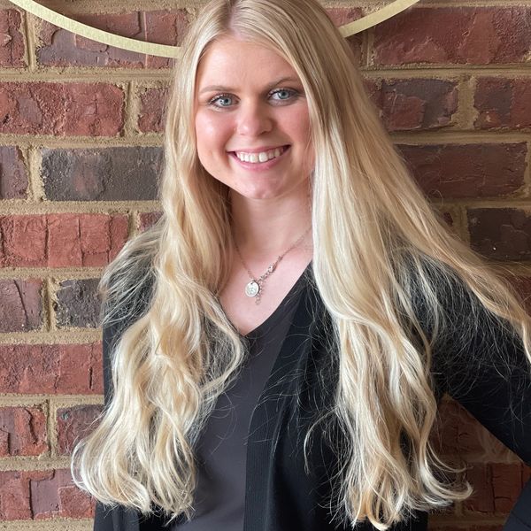 Smiling young woman with long blonde hair stands against a brick wall.