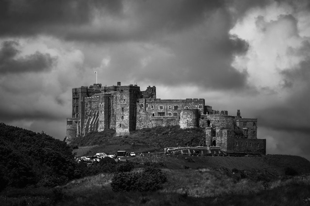 A black and white photo of an imposing Bamburgh Castle atop its rolling hill home with moody clouds.