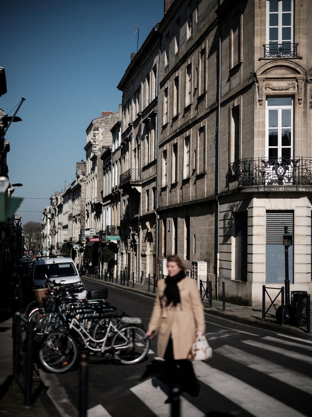 A vertical colour photograph showing a woman, out of focus, crossing a road in Bordeaux, France. 