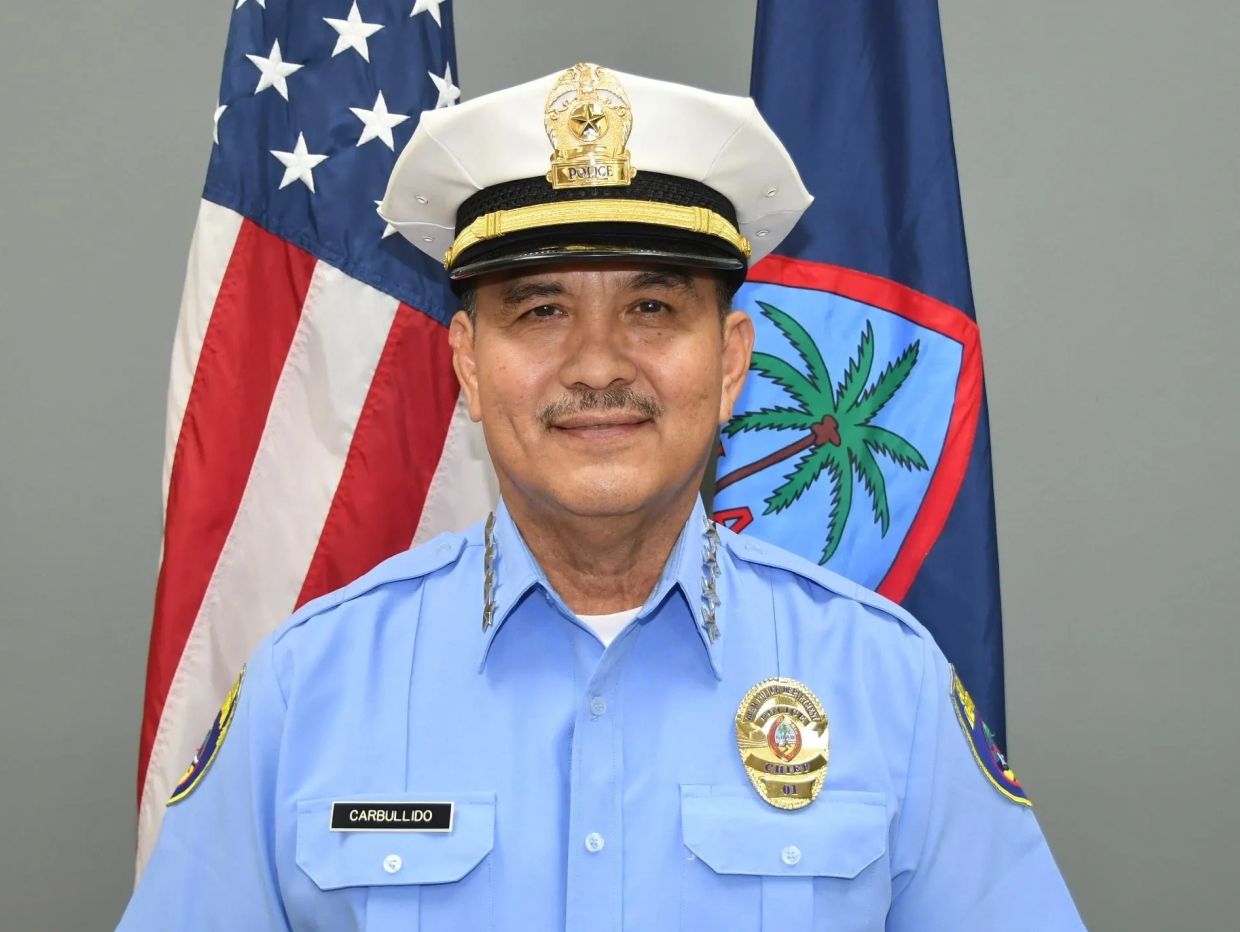 Police officer in uniform standing in front of American and Guam flags.