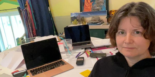 Woman sitting at a cluttered desk with two laptops and office supplies.
