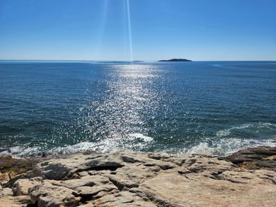 Sunlight glistens on calm ocean waters near rocky shore with distant island.