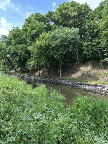 A peaceful creek bordered by green trees and grassy banks under a blue sky.