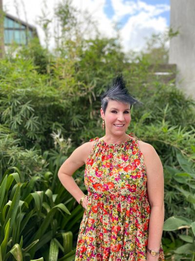 Woman in a floral dress standing in a lush garden, smiling.