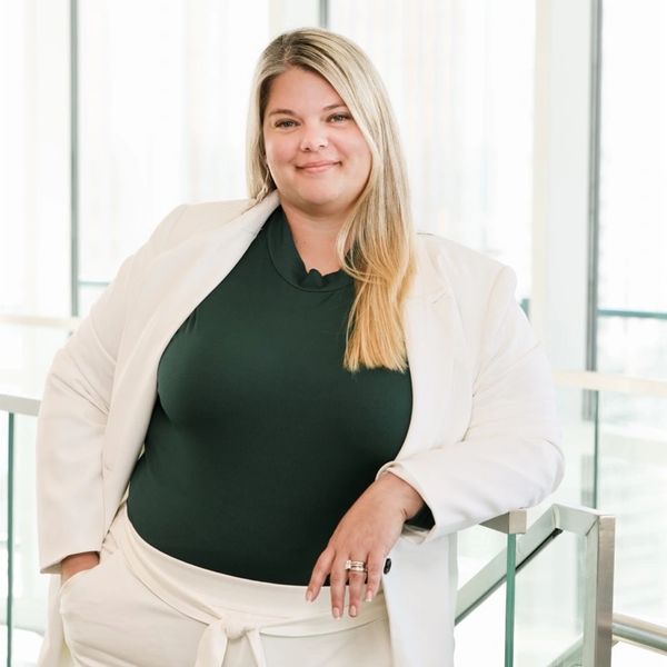 Confident woman in white suit and dark top leans on railing indoors.