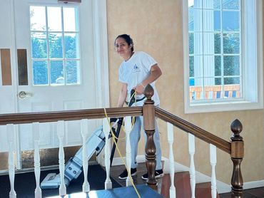 Woman vacuuming the floor near a staircase inside a bright room.