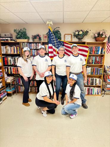 Group of six people in matching shirts and caps posing indoors with an American flag.