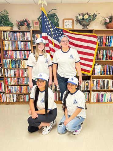 Four girls in matching shirts and caps pose in front of an American flag inside a library.