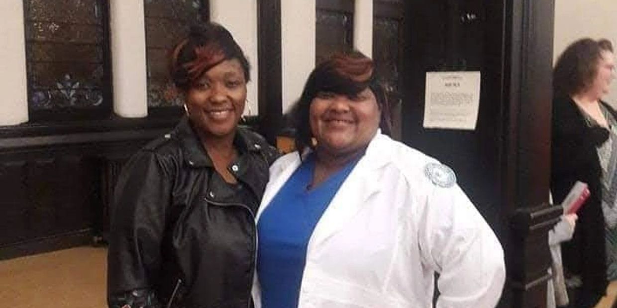 Two women smiling indoors, one in a black jacket and the other in a white medical coat.