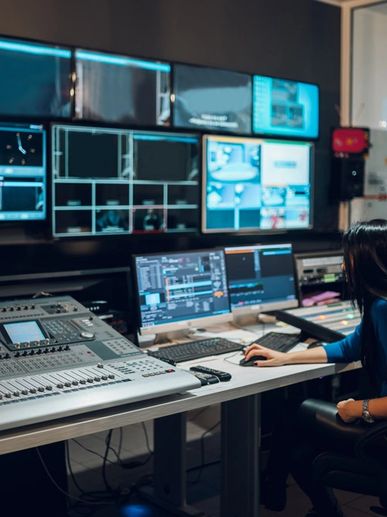 Person working at a media control room with multiple screens and audio equipment.