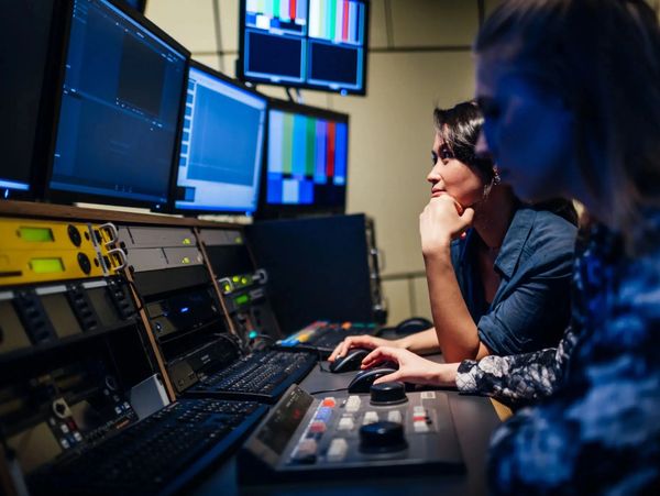 Two women working at a video editing control room with multiple monitors and equipment.